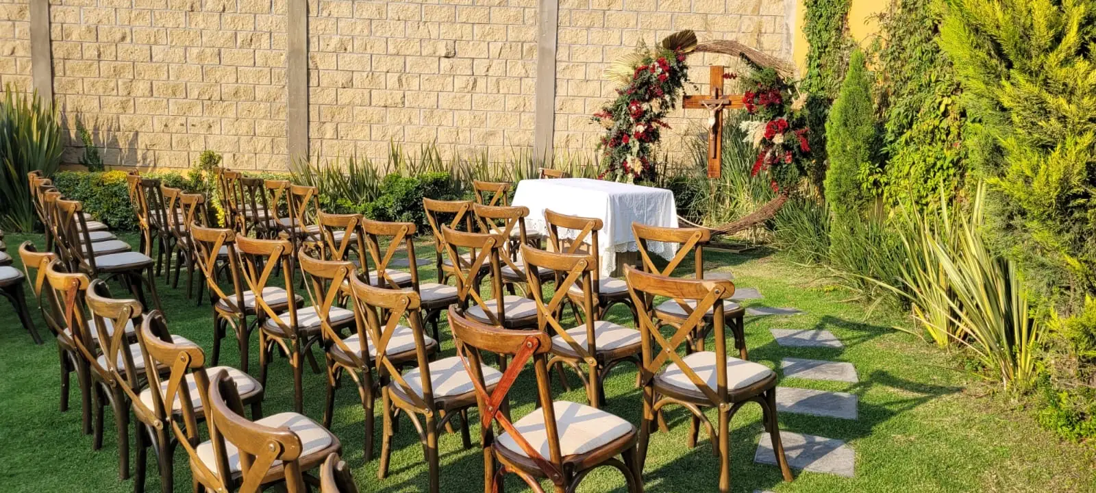 Cruz con arco de flores rojas y mesa de altar en el jardín de Bonanza, con sillas de madera bajo la luz dorada de la tarde