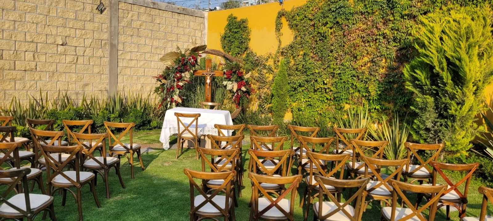 Jardín de Bonanza con luz dorada de la tarde: arco floral con cruz, mesa de altar y sillas crossback
