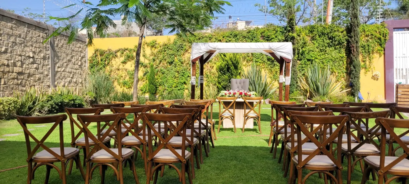 Pérgola blanca del jardín de Bonanza entre muros de enredadera, con sillas de madera listas para la ceremonia