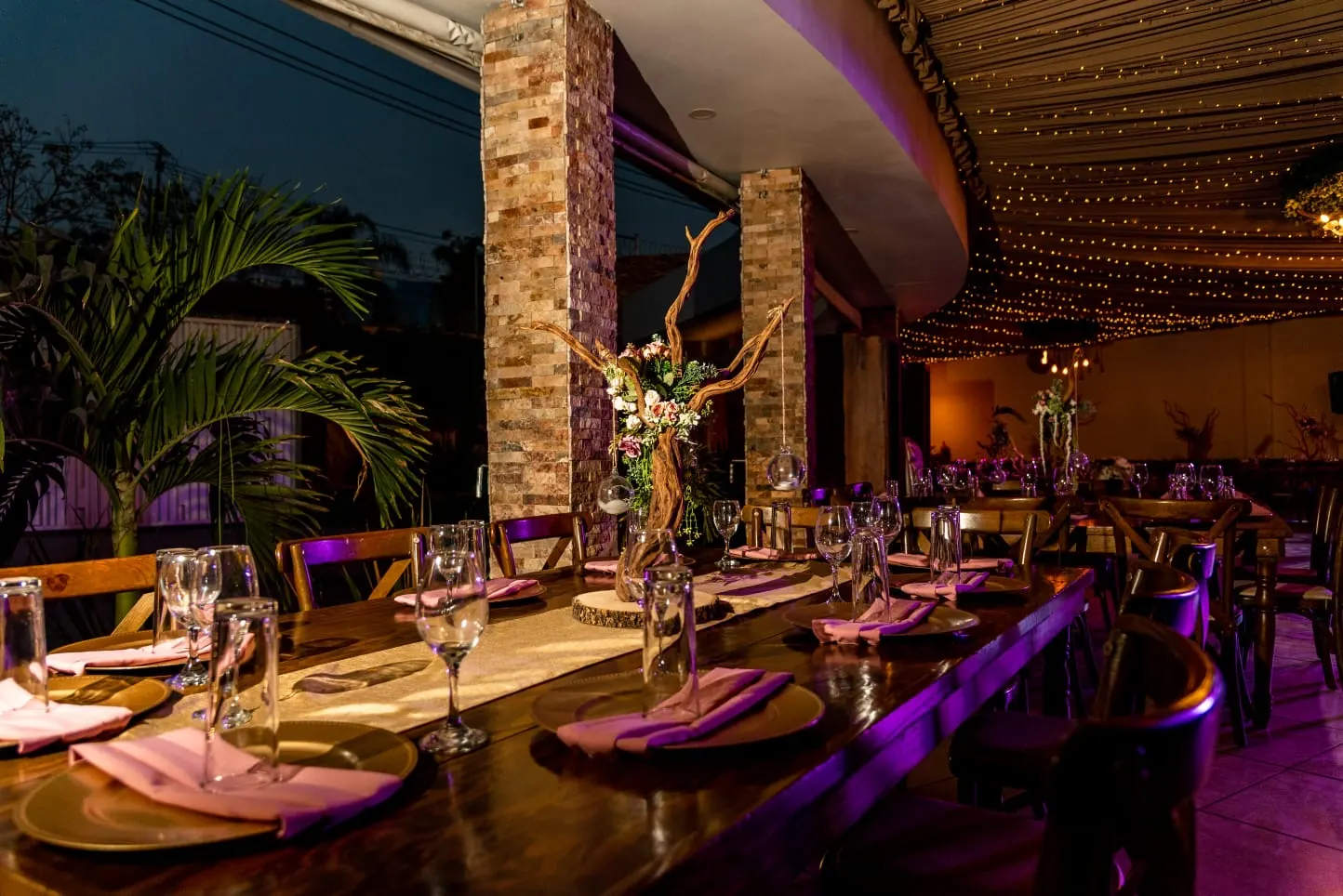 Terraza de Bonanza de noche: mesa larga de madera con servilletas rosas bajo el cielo de luces cálidas, junto a la palmera